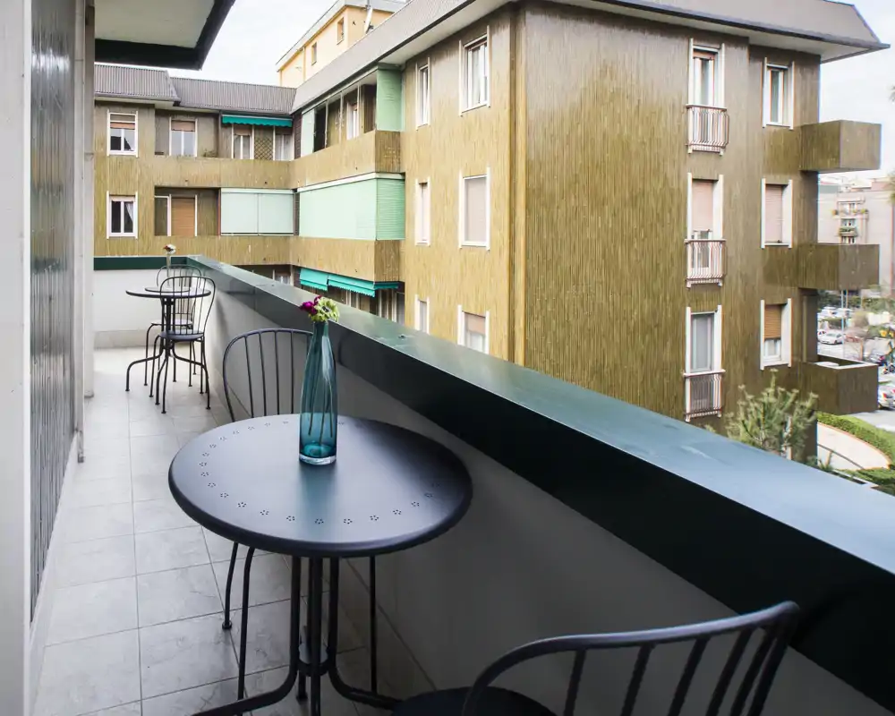 Ampio balcone privato con tavolini e sedie in metallo stile bistro, pavimento piastrellato e vista sugli edifici vicini — ben arredato con un vaso decorativo.