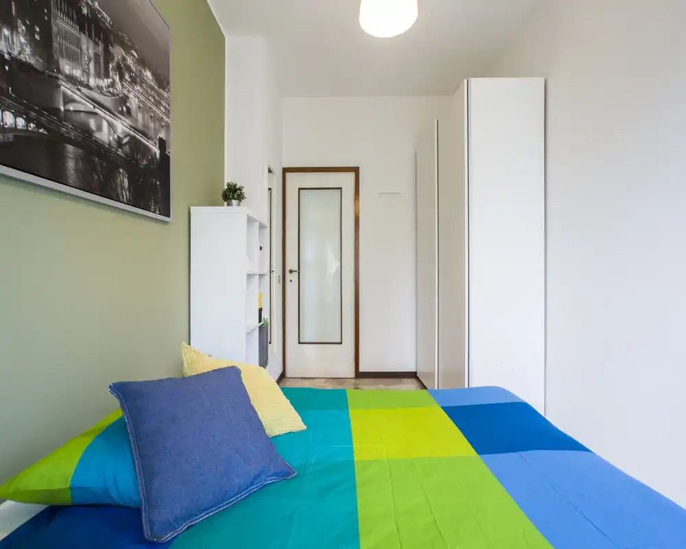 Camera da letto luminosa e moderna con una trapunta colorata, due cuscini decorativi e armadi bianchi. L'immagine mostra un letto ben illuminato in primo piano con guardaroba e mensole lungo la parete.