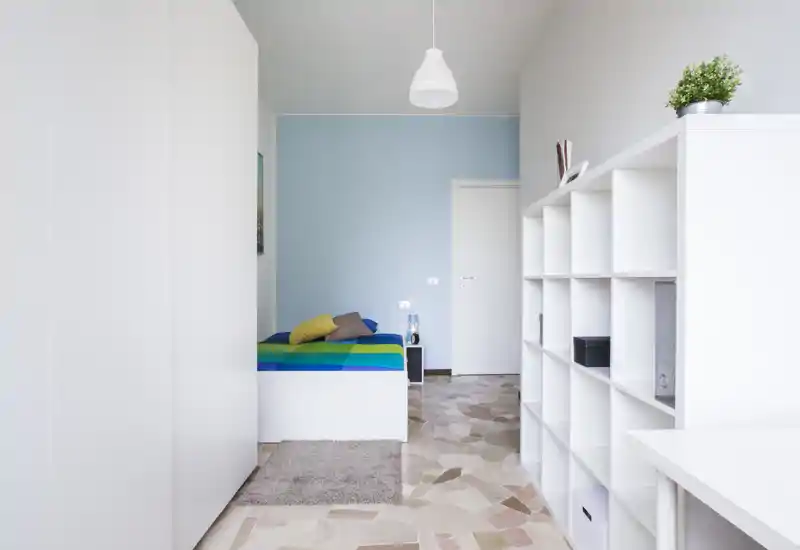 Camera da letto luminosa e moderna con un letto singolo ben rifatto contro una parete azzurro chiaro, mobili contenitori bianchi e uno stile minimalista adatto agli affitti brevi.