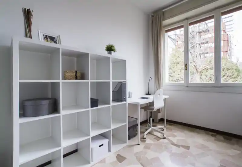 Zona giorno/studio luminosa e minimal con ampia finestra, scaffale bianco e scrivania compatta—ideale per lavoro o lettura.