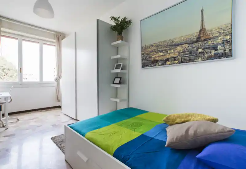 Camera da letto singola moderna e luminosa con letto colorato, ampia finestra che illumina la stanza, armadio e decor minimal con quadro di Parigi.