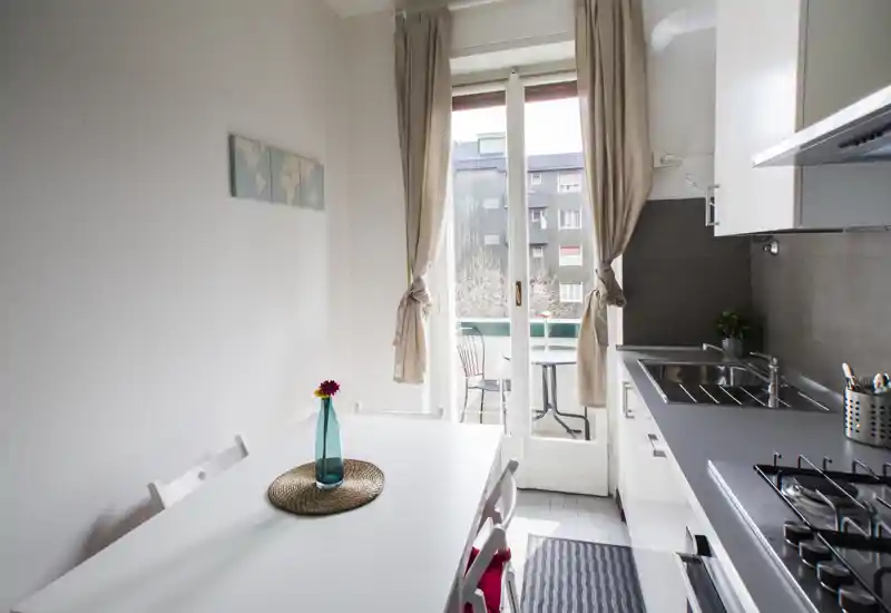 Cucina compatta e luminosa con tavolo da pranzo e accesso diretto a un piccolo balcone. Mobile cucina moderno, piano cottura a gas, lavello in acciaio e un vaso decorativo che rendono lo spazio ordinato e funzionale per un appartamento cittadino.