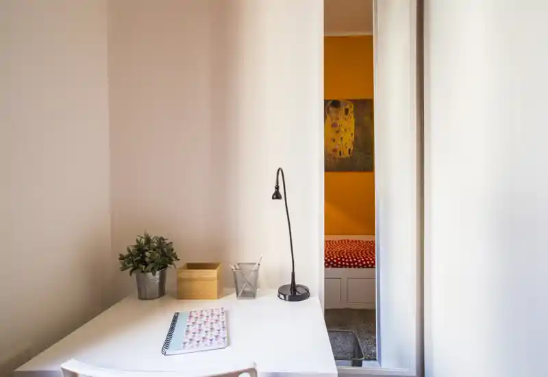 Cosy bedroom corner seen through a doorway with a small table and lamp in the foreground; the bed with patterned bedding is visible in the adjacent room.