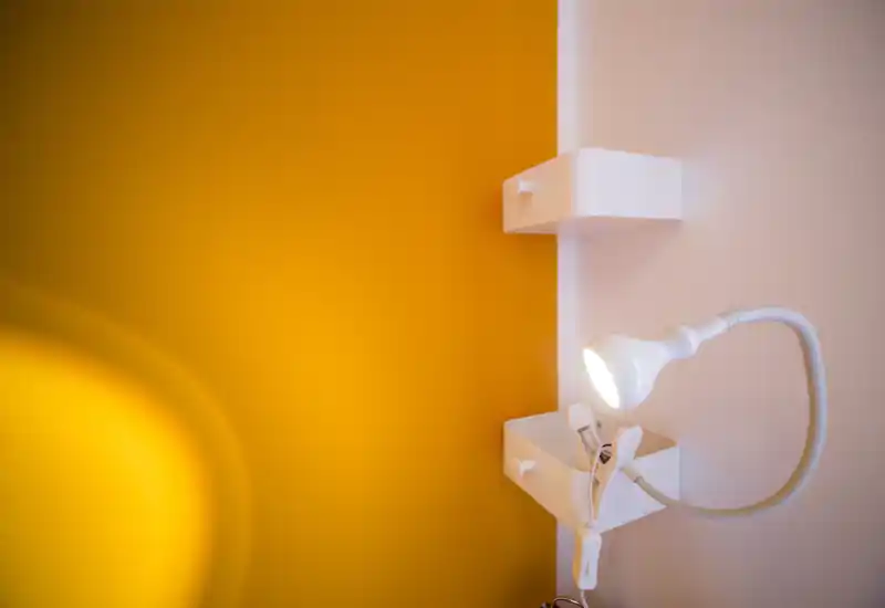 Close-up of a warm, modern wall corner with a flexible reading lamp and small white wall-mounted storage boxes; vivid yellow paint creates a bright, contemporary accent.