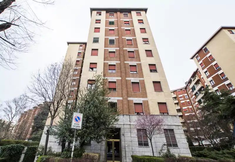 Exterior view of a residential apartment building with multiple floors, entrance door and landscaped greenery in front — suitable as an outdoor/building facade shot for a listing.