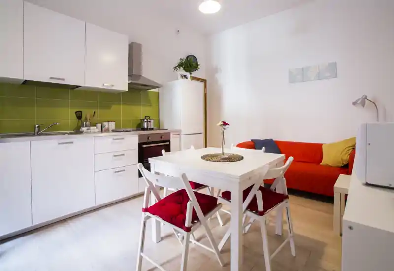 Bright, modern open-plan kitchen with white cabinets, green tile backsplash, a dining table with red-cushioned chairs and a cozy red sofa — ideal for small apartment listings.
