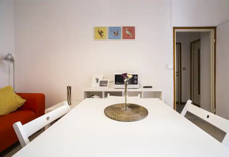 Bright, modern living/dining area with a white table, decorative centerpiece and a red sofa with cushions; clean minimalist styling and soft natural light.