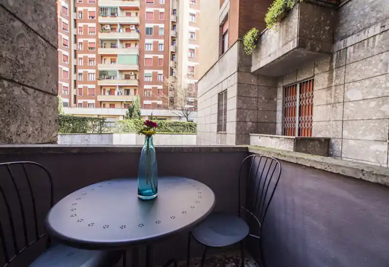 Cozy urban balcony with a small round table, two metal chairs and a decorative vase — ideal for morning coffee while overlooking surrounding apartment buildings.