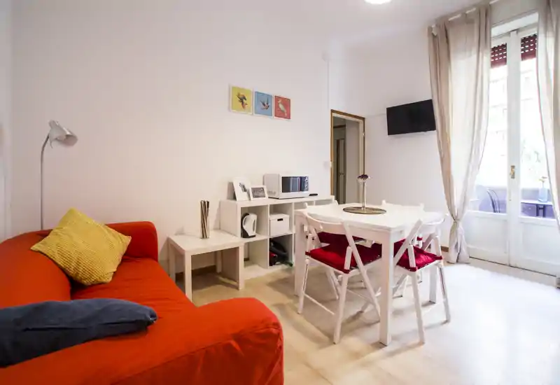 Bright, well-staged living room with a red sofa, dining table for four and large window letting in natural light — cozy and modern space ideal for relaxing or entertaining.