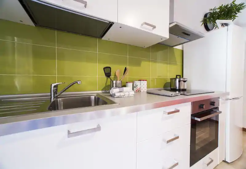 Bright modern kitchen with stainless steel countertop, integrated sink, electric oven and a white fridge, highlighted by a green tile backsplash and neat utensil arrangement.