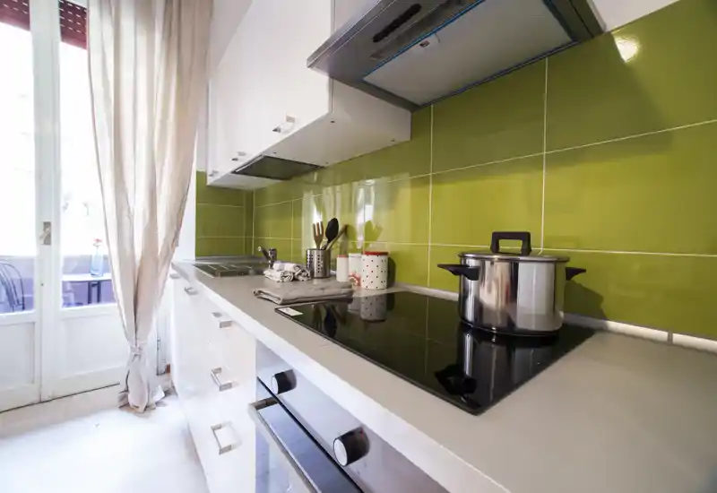 Bright modern kitchen with a sleek induction hob, stainless pot, and green tiled backsplash; large glass door with light and a soft curtain adds warmth.