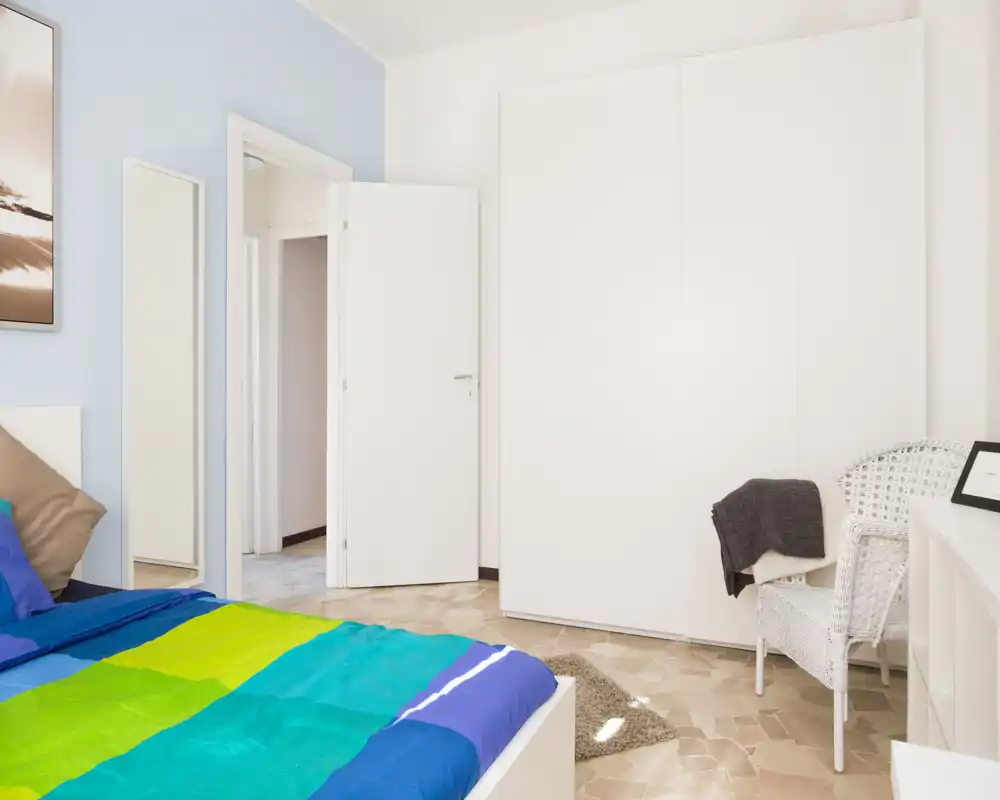 Dormitorio luminoso y bien presentado con colcha colorida, amplio armario blanco y una silla de mimbre — ideal para una estancia acogedora.