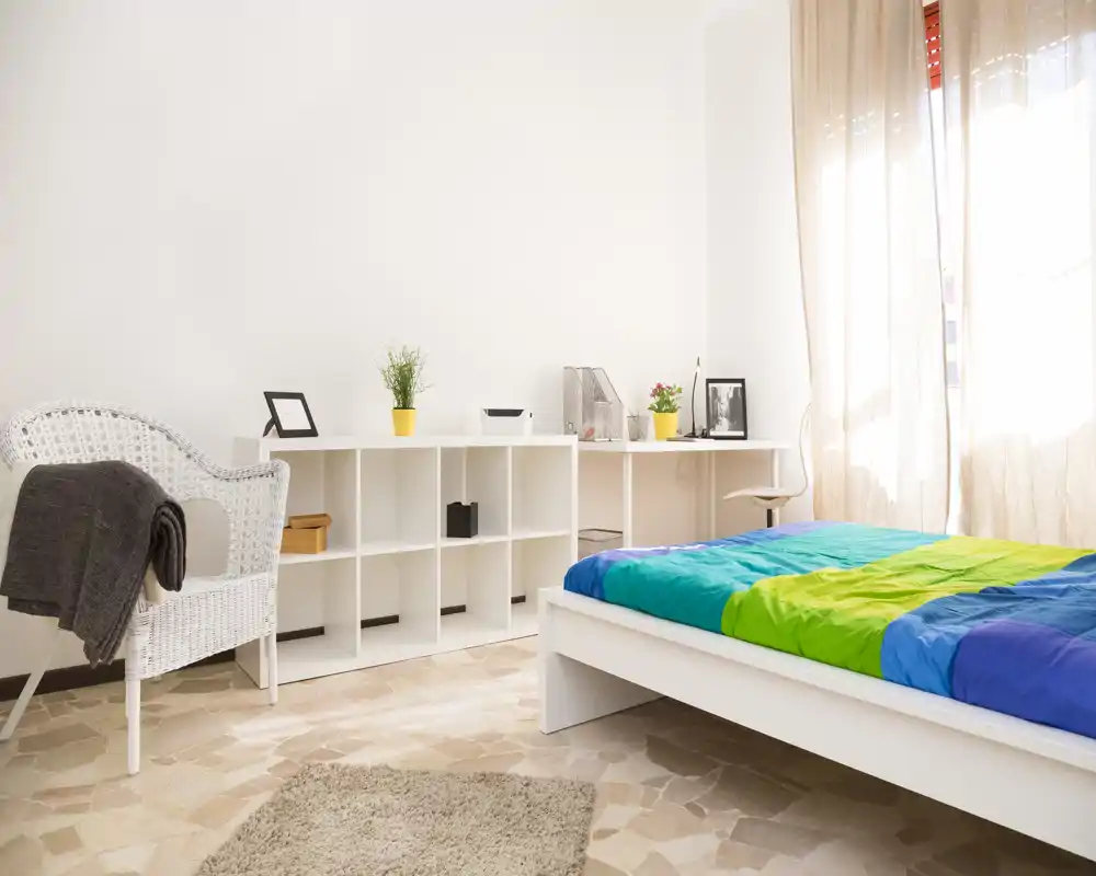 Dormitorio moderno y luminoso con cama baja blanca y ropa de cama colorida, sillón de mimbre, estantería y una ventana soleada con cortinas ligeras.