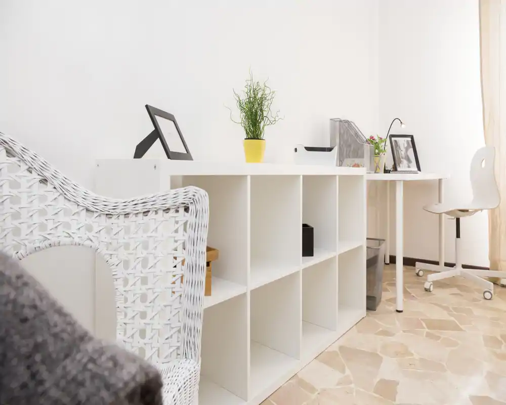 Zona de estar luminosa y minimalista con módulo de almacenamiento blanco, sillón de mimbre y un pequeño escritorio — espaciosa, sencilla y bien iluminada.