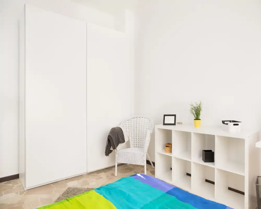 Dormitorio luminoso y minimalista con colcha colorida, armario blanco y estantería; estilo sencillo y fresco, ideal para un anuncio de alquiler.
