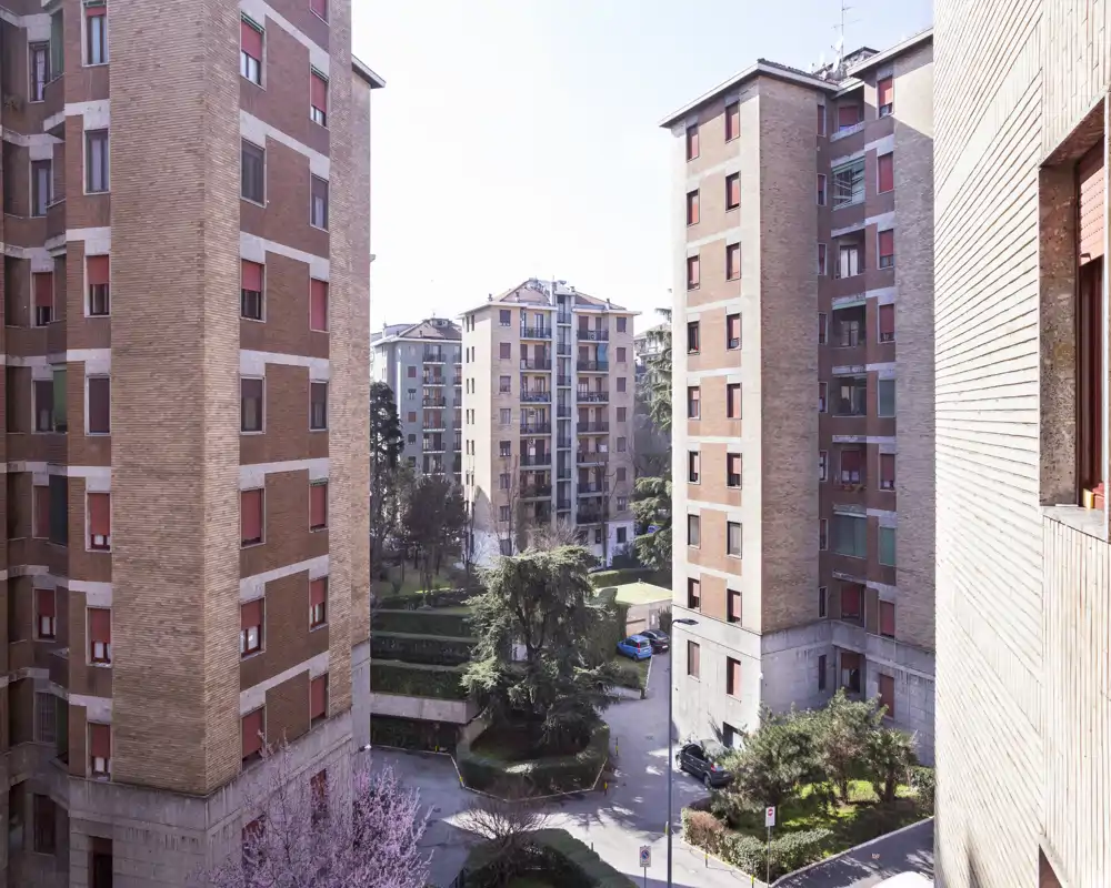 Vista luminosa del patio interior enmarcado por edificios de ladrillo, con árboles, caminos y coches aparcados — ideal para mostrar el espacio exterior común.