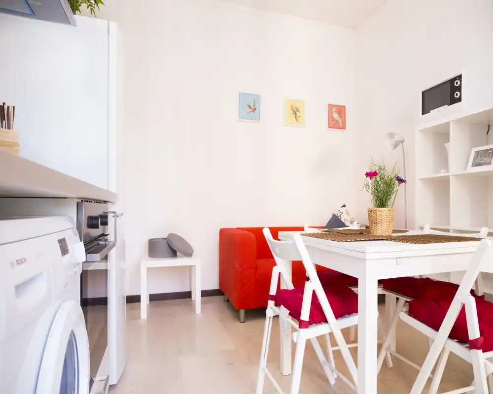 Zona de estar luminosa y compacta con un sofá rojo, mesa y sillas blancas, y electrodomésticos de cocina visibles en el borde — ideal para un estudio o apartamento pequeño.