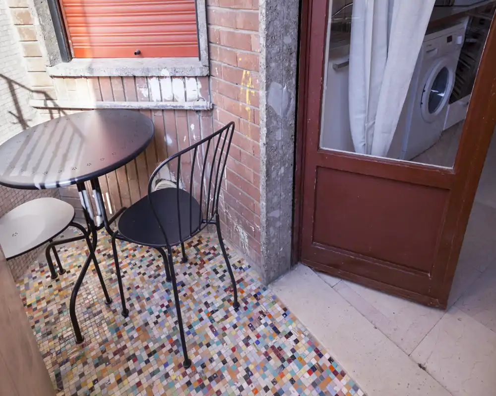 Pequeño balcón enlosado con una mesa redonda de metal y dos sillas, suelo de mosaico colorido y vista a una zona de lavado interior a través de la puerta de cristal.