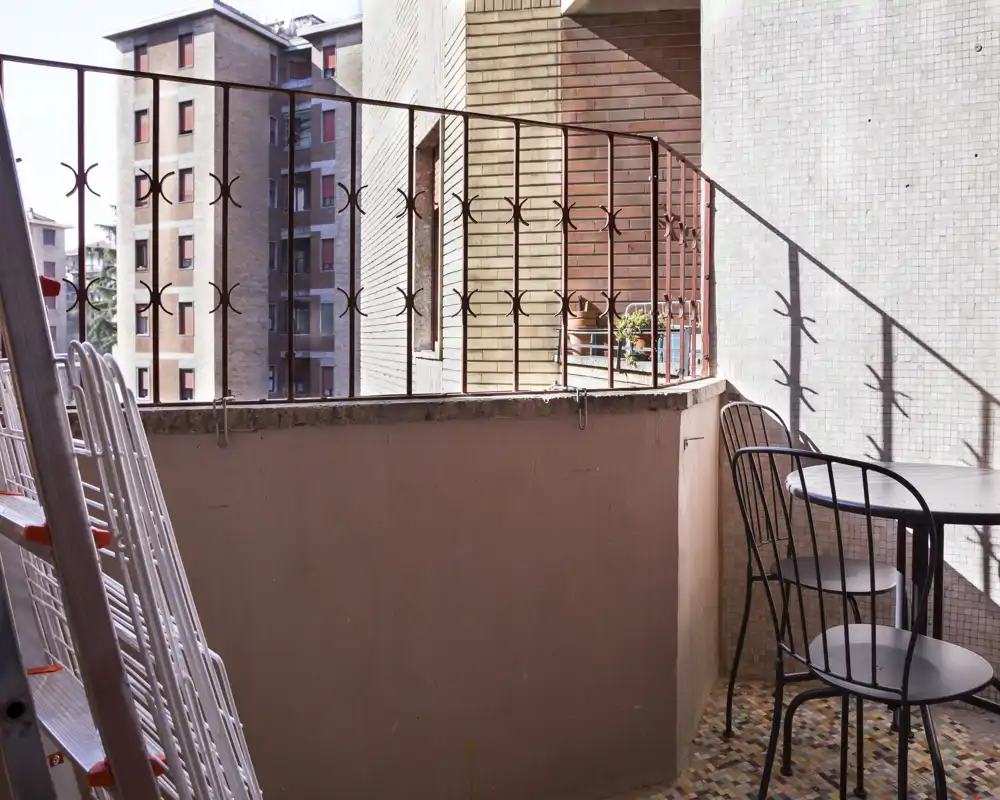 Pequeño balcón soleado con barandilla de metal, suelo de mosaico y una mesa redonda con dos sillas — ideal para tomar un café por la mañana o una pausa al aire libre.