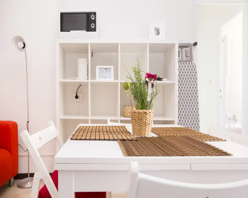 Zona de comedor luminosa y bien arreglada con mesa blanca, manteles individuales de mimbre y una planta como centro; estantería y microondas al fondo indican una cocina abierta.
