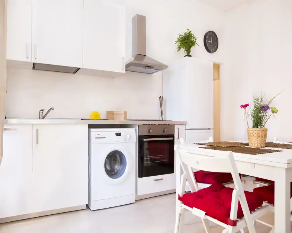 Cocina moderna y luminosa con armarios blancos, horno empotrado y lavadora, además de una mesa de comedor con sillas con cojines rojos y plantas decorativas.