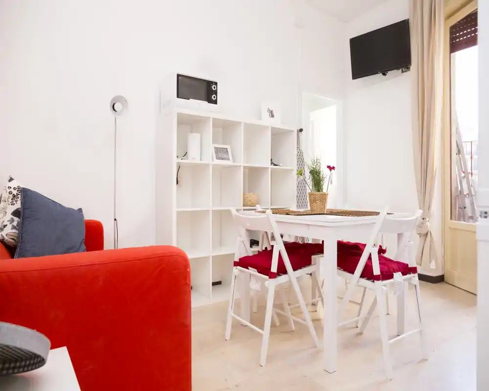 Zona de estar/comedor luminosa y moderna con un sofá rojo, mesa blanca y sillas plegables con cojines rojos; estantería minimalista y mucha luz natural desde la puerta.