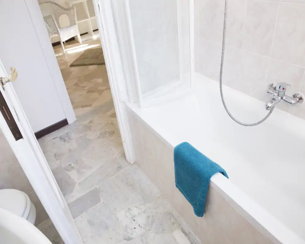 Baño limpio y bien iluminado con bañera y ducha de mano, azulejos blancos y una toalla turquesa colgada del borde de la bañera.