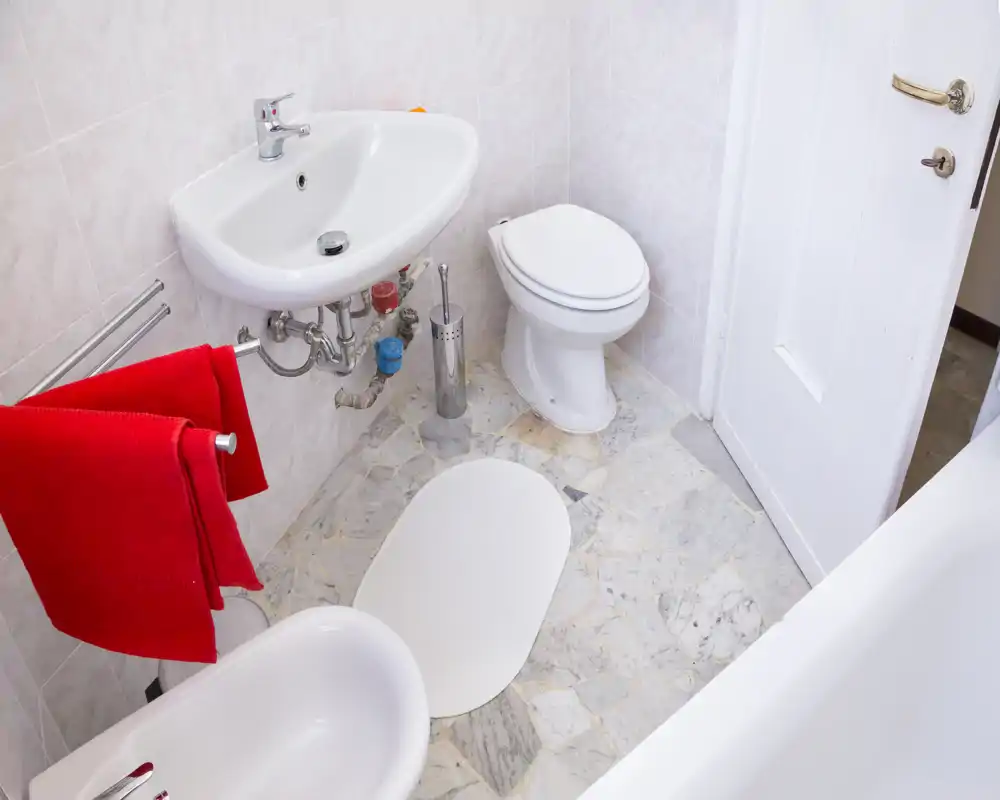 Baño pequeño y limpio con lavabo de pared, inodoro, bidé y borde de bañera visible; toallas rojas añaden un toque de color.