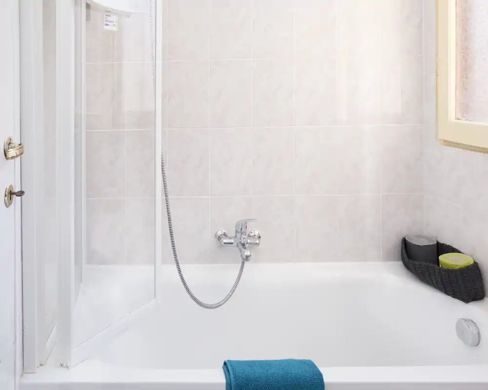 Baño limpio y luminoso con bañera, grifo mural y ducha de mano; azulejos claros y una toalla sobre el borde que aportan una sensación fresca.