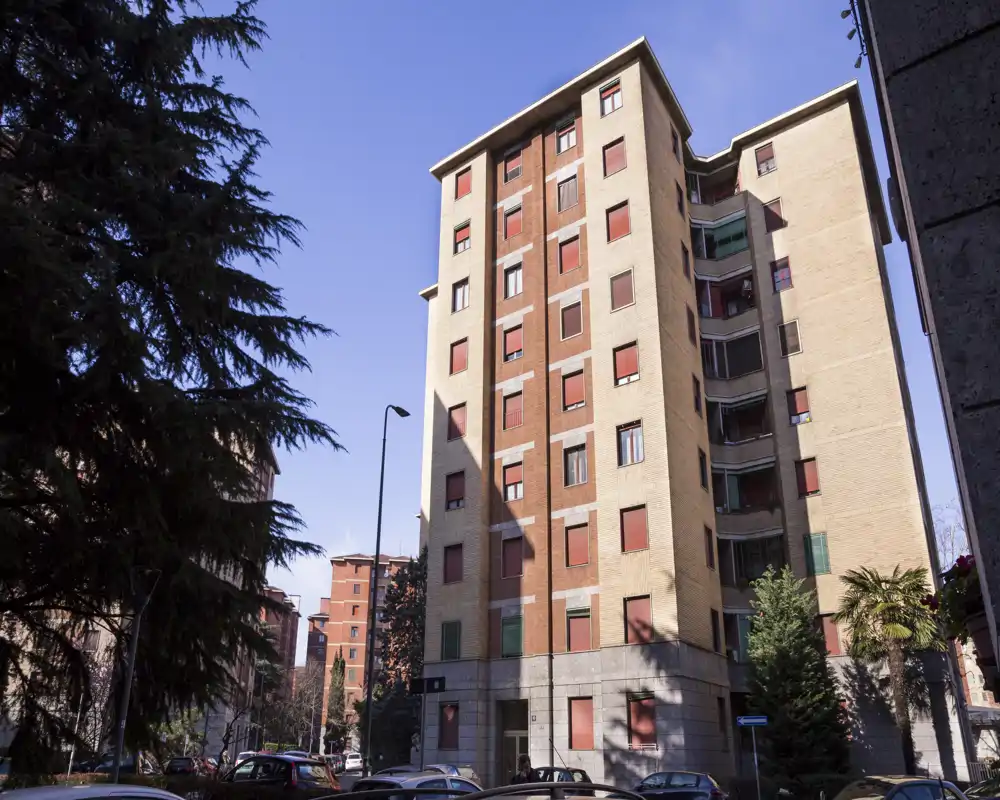Vista exterior de un edificio de apartamentos residenciales a plena luz del día, mostrando las fachadas, ventanas y la calle con coches aparcados y árboles.