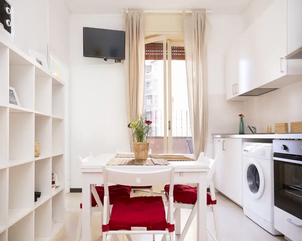 Cocina compacta y luminosa con mobiliario blanco, horno y lavadora integrados, y una pequeña mesa con sillas con cojines rojos frente a la puerta del balcón.