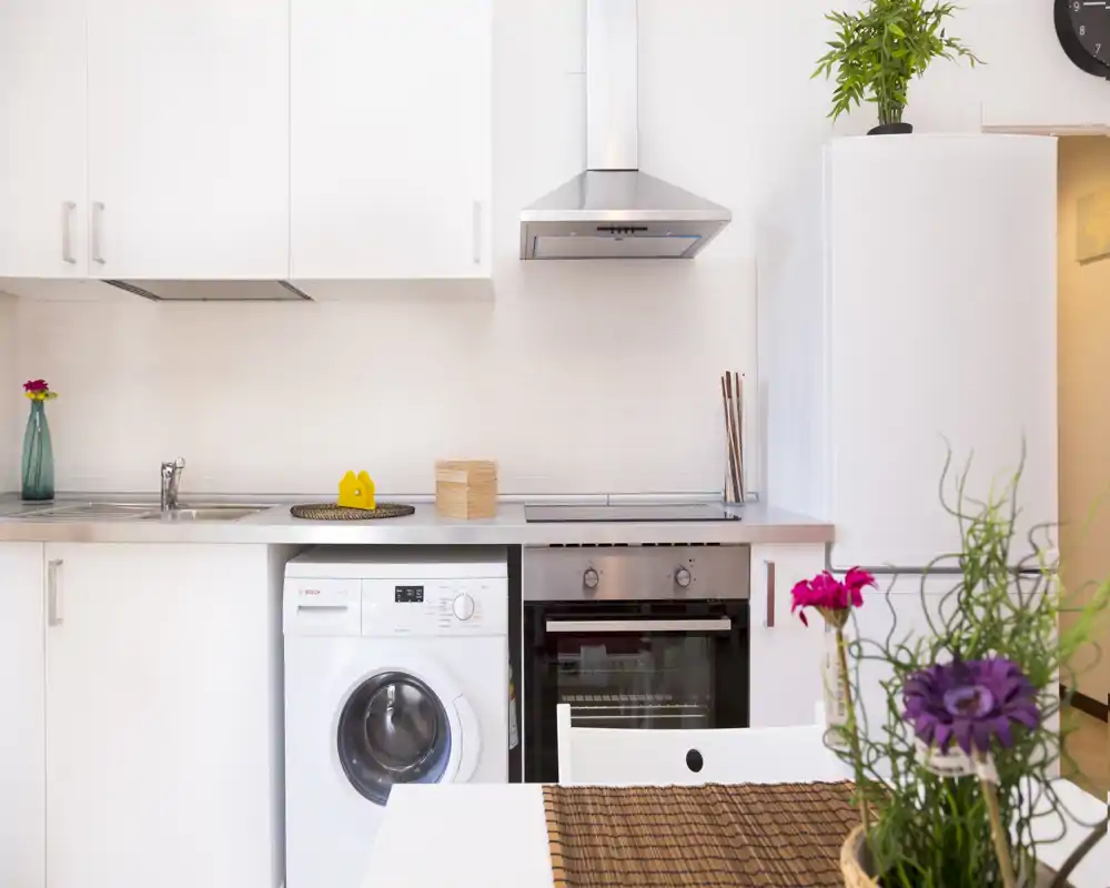 Cocina moderna y compacta con muebles blancos, horno empotrado, lavadora y nevera; diseño luminoso y limpio con plantas decorativas.