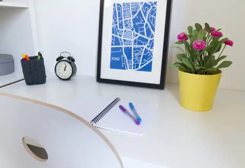 Rincón de trabajo luminoso y ordenado con escritorio blanco, mapa enmarcado, despertador y planta en maceta amarilla — perfecto para trabajar o estudiar.