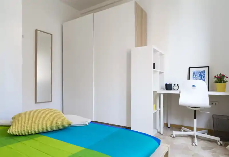 Dormitorio moderno y luminoso con colcha colorida, armario alto y una zona de trabajo compacta, ideal para estudiantes o profesionales.