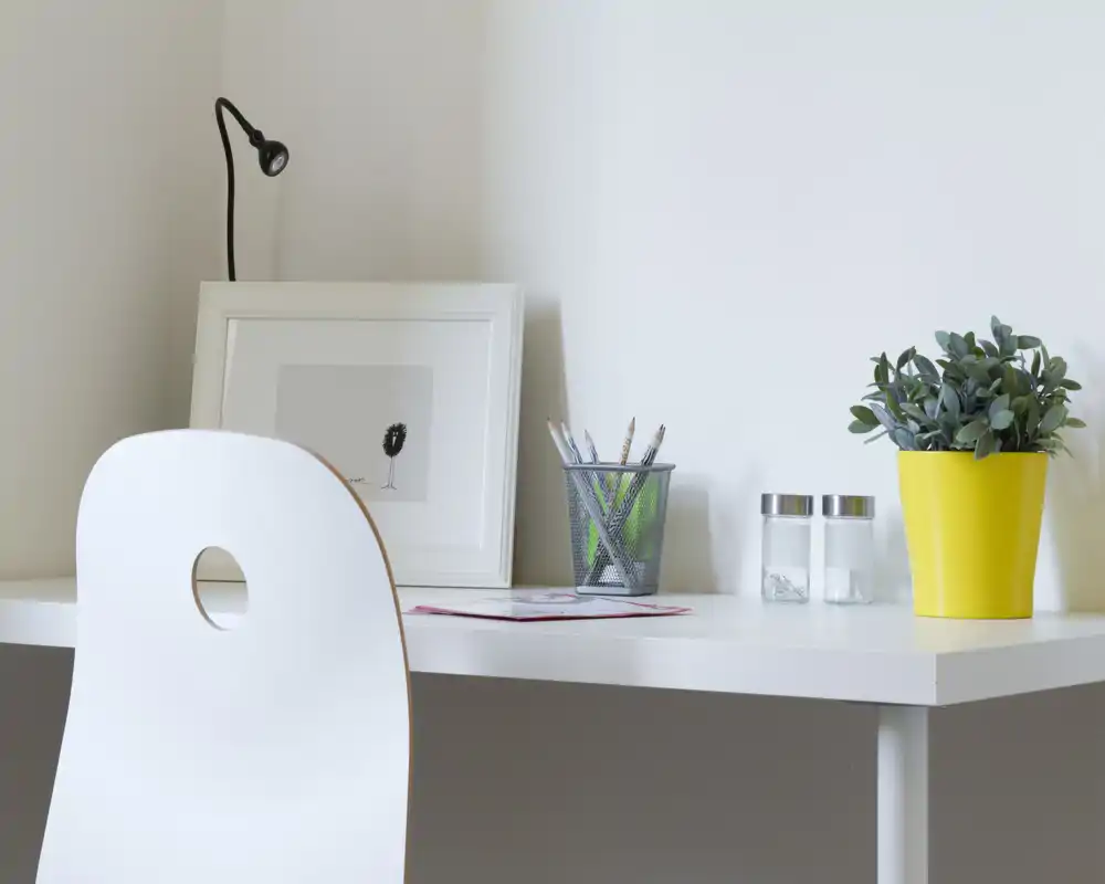 Angolo scrivania minimalista e ordinato con sedia bianca, stampa incorniciata, portamatite e una pianta in vaso — un piccolo spazio di lavoro luminoso in un'area giorno.