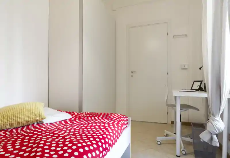 Dormitorio moderno y luminoso con colcha roja de lunares y un pequeño escritorio junto a la ventana. Espacio funcional y bien iluminado.