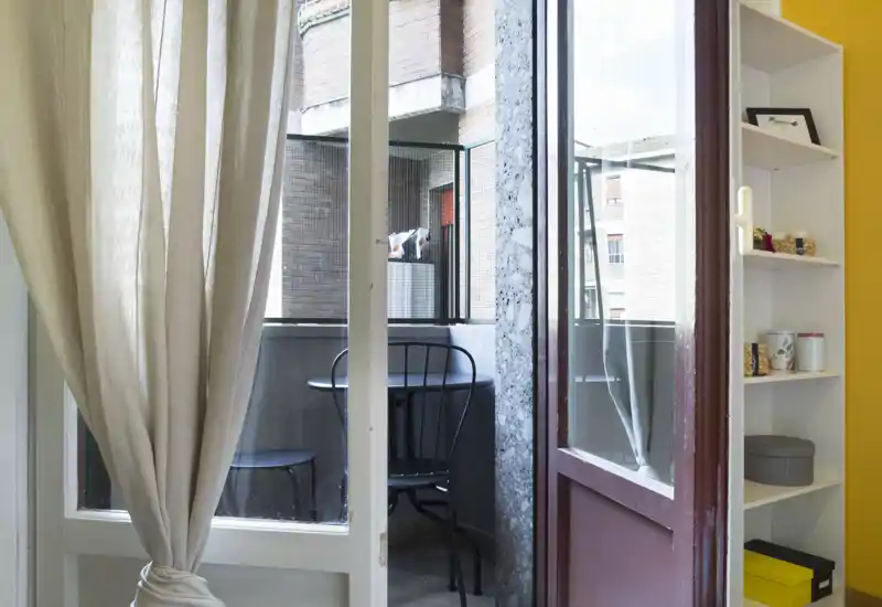 Pequeño balcón privado con mesa redonda de metal y dos sillas, visto a través de una puerta con cortinas; espacio exterior luminoso para comer al aire libre.