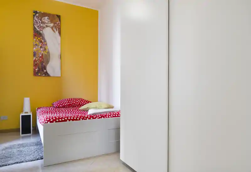 Dormitorio moderno y compacto con cama blanca, ropa de cama roja con lunares, pared de acento amarilla y una mesita estrecha; espacio luminoso y bien compuesto, ideal para una persona.