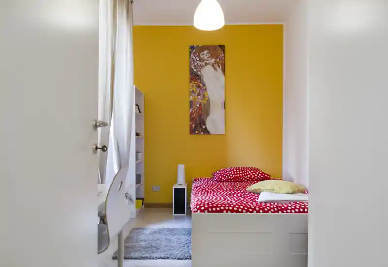 Dormitorio luminoso y compacto con cama individual blanca con ropa de cama roja de lunares, alfombra suave, estantería y una pared amarilla con cuadro — bien iluminado y acogedor.