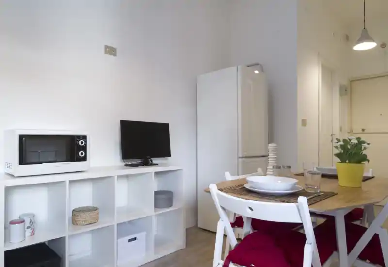 Cocina abierta y compacta con nevera, microondas y una pequeña mesa de madera para dos. Luminoso, minimalista y bien decorado — ideal para estancias cortas.
