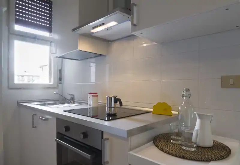Cocina moderna y compacta con placa de inducción, horno integrado, fregadero de acero inoxidable y accesorios ordenados. La luz de la ventana y el alicatado blanco crean un espacio limpio y funcional, ideal para alquileres de corta estancia.