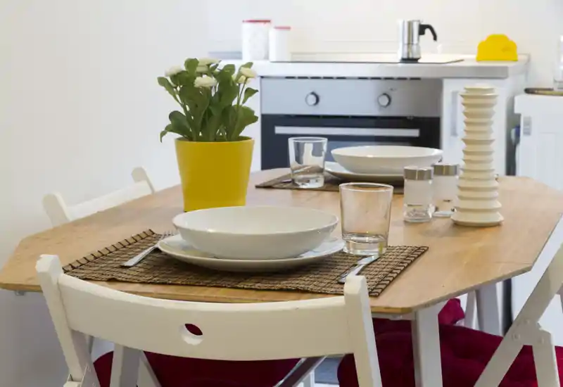 Mesa de comedor luminosa y bien puesta con platos, vasos y una planta decorativa en un área cocina-comedor compacta — ideal para comidas en grupo pequeño.