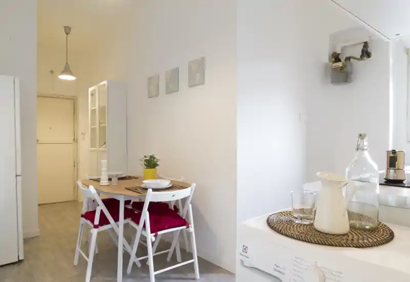 Acogedor rincón de comedor luminoso con mesa de madera, cuatro sillas plegables blancas con cojines rojos, vajilla sencilla y una pequeña planta — ideal para comidas en un apartamento pequeño.