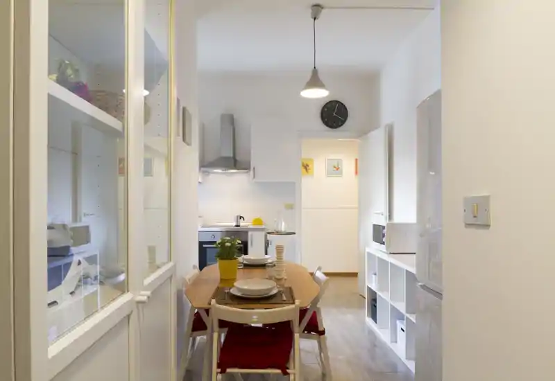 Zona de comedor luminosa y cuidada con mesa de madera para cuatro, cojines rojos y cocina visible al fondo — ideal para anuncios de apartamentos pequeños.