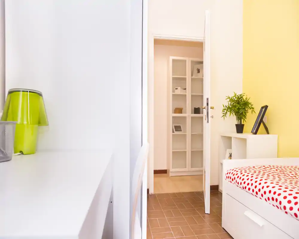 Dormitorio luminoso y minimalista con una cama individual con edredón de lunares rojos, una mesita con planta y marco, y una zona de escritorio blanca y limpia. La habitación es aireada y de decoración sencilla, ideal para una persona o estancias cortas.