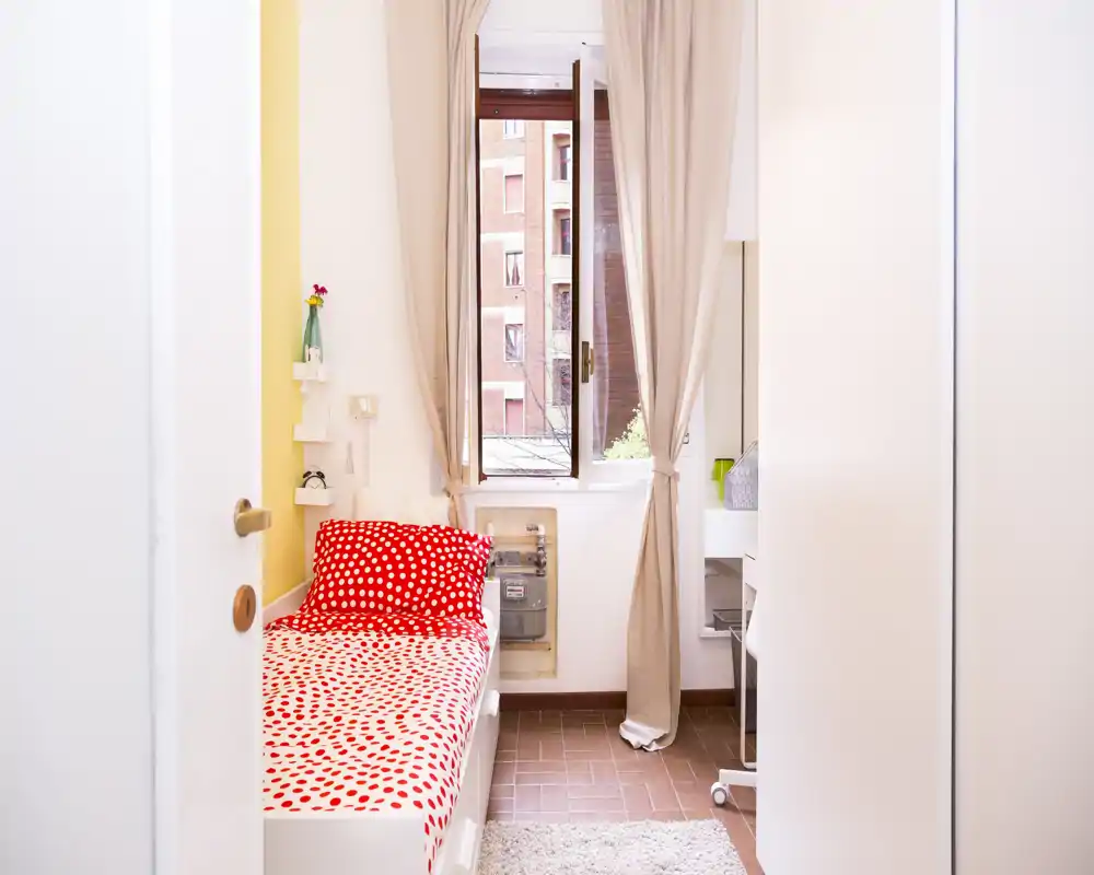 Dormitorio individual luminoso con cama roja de lunares blancos, luz natural desde una ventana con cortinas beige y una zona de escritorio compacta — acogedor y bien decorado para estudiante o un inquilino solo.