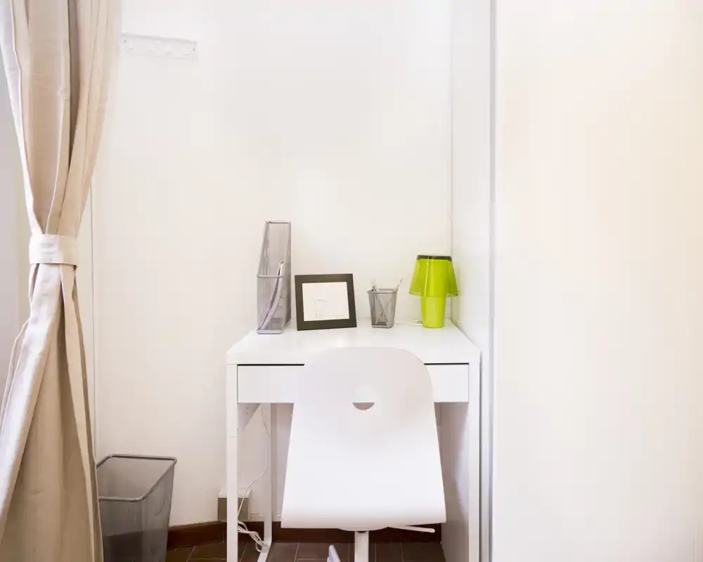 Pequeño y ordenado espacio de trabajo con escritorio blanco, silla a juego y lámpara verde; cortinas neutras y decoración mínima crean un rincón funcional y tranquilo, ideal para teletrabajo.