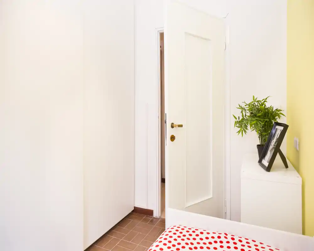 Rincón de dormitorio luminoso y minimalista con la cama parcialmente visible en primer plano, un armario blanco, un mueble pequeño con una planta y una foto enmarcada y una puerta abierta al pasillo.