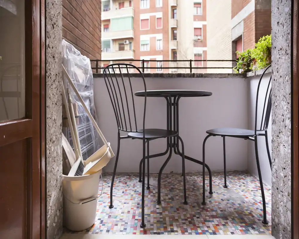 Pequeño balcón en la ciudad con mesa redonda de metal y dos sillas, suelo de mosaico colorido y vista a edificios vecinos — ideal para el café de la mañana.