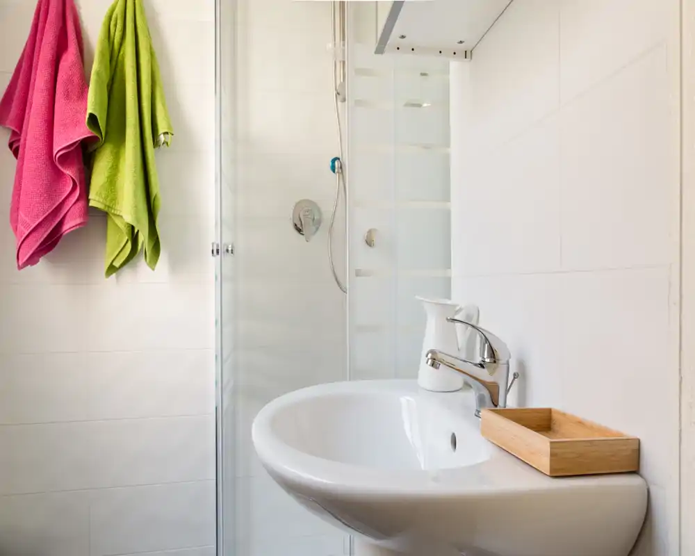Baño compacto y luminoso con lavabo blanco suspendido, grifo cromado moderno, ducha con mampara de cristal y toallas de colores que aportan contraste.